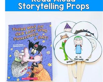以下が含まれることがあります： イラスト付きの「THERE WAS AN OLD LADY WHO SWALLOWED A BAT！」というタイトルの子供向けの本と、棒の上のいくつかの丸いキャラクター小道具。小道具には、ゴブリン、おばあさん、その他のキャラクターが含まれます。「Read Aloud Storytelling Props」というテキストが上部にあります。