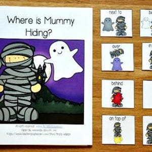 May include: A spiral-bound children's book titled "Where is Mummy Hiding?" with illustrations of a mummy and ghost. Accompanying cards show positional words like "next to," "between," and "under," with corresponding mummy and ghost graphics. The background is a wooden surface.