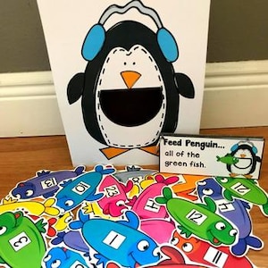 May include: A white box featuring a cartoon penguin wearing blue earmuffs and a large open mouth. Brightly coloured fish cutouts with numbers and letters are scattered in front. A small card reads "Feed Penguin... all of the green fish."