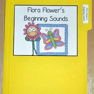 May include: A yellow file folder with a white label displaying the text "Flora Flower's Beginning Sounds." The label features a cartoon flower and a butterfly. A smaller tab label reads "Flora Flower's Beginning Sounds."