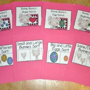 May include: Pink file folders with educational printouts featuring a cartoon bunny. The printouts include titles like "Counting Eggs," "Shape Match," and "Beginning Sounds." The folders are arranged on a light-colored surface.