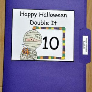 以下が含まれることがあります： 「Happy Halloween Double It」の文字とミイラのイラスト、カボチャを持つ白い長方形が特徴の紫色のファイルフォルダー。数字の10が大きく表示されています。