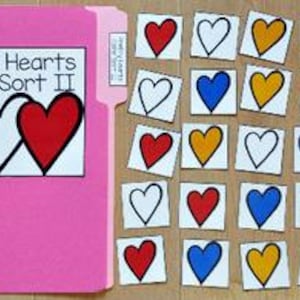 Op de afbeelding: Een roze map met de tekst "Pretty Hearts Color Sort II" en verschillende hartvormige kaarten. De kaarten tonen contouren van harten in verschillende kleuren, waaronder blauw, wit, rood en geel. De kaarten liggen verspreid op een lichtgekleurd houten oppervlak.