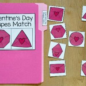 以下が含まれることがあります： 「Valentine's Day Shapes Match」と書かれたピンク色のファイルフォルダーと、ピンク色の封筒と赤いハートが描かれたマッチングシェイプカード。カードは木製の表面に散らばっており、さまざまな幾何学的な形を示しています。