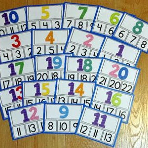 Może przedstawiać: Zestaw kolorowych kart z liczbami, każda z liczbą od 1 do 20. Karty mają niebieską obwódkę, białe tło i ciąg liczb poniżej. Karty są ułożone na drewnianej powierzchni.