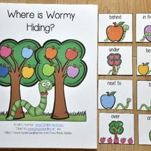 May include: A spiral-bound children's book titled "Where is Wormy Hiding?" with illustrations of trees, apples, and a worm. Accompanying cards show prepositions like "behind," "in front of," and "on top of."