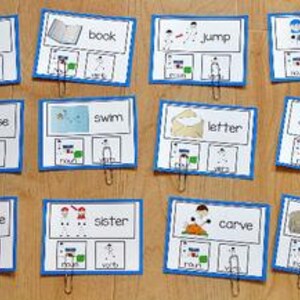 Puede incluir: Doce tarjetas didácticas con bordes azules y clips metálicos están dispuestas sobre una superficie de madera. Cada tarjeta presenta una palabra, una ilustración y etiquetas para sustantivo, verbo y pronombre. Palabras: "cavar", "libro" y "saltar".