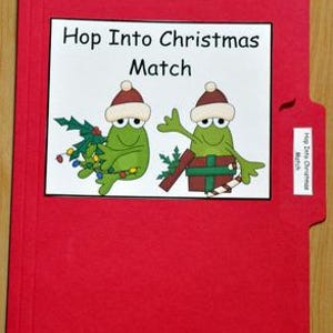 以下が含まれることがあります： 「Hop Into Christmas Match」の文字が入った白いラベルが付いた赤いファイルフォルダー。ラベルには、サンタ帽をかぶった2匹の漫画のカエルが描かれており、1匹はクリスマスツリーを、もう1匹はプレゼントを持っています。