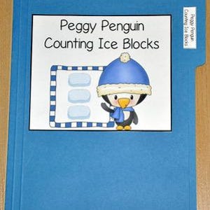May include: A blue file folder with a white label featuring a cartoon penguin wearing a blue hat and scarf. The text "Peggy Penguin Counting Ice Blocks" is above the penguin. A smaller label on the tab reads "Peggy Penguin Counting Ice Blocks".