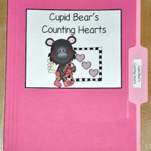 以下が含まれることがあります： 「Cupid Bear's Counting Hearts」のテキストが書かれた白いラベルが付いたピンク色のファイルフォルダー。ラベルには、ハートと3つのピンクのハートを持ったクマのイラストが含まれています。ファイルフォルダーには同じテキストのタブが付いています。