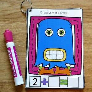 Pode incluir: Um livro de atividades educativas colorido com uma ilustração de monstro azul e o texto "Draw 2 More Eyes." Uma caneta de quadro branco rosa está ao lado do livro. O livro tem um anel de metal na parte superior.