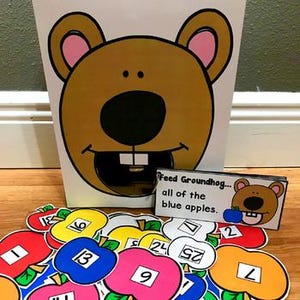 Puede incluir: Un recipiente blanco con una cara de marmota de dibujos animados, con una abertura para la boca. Recortes de manzanas coloridas con números están esparcidos al frente. Una pequeña tarjeta dice "Feed Groundhog... all of the blue apples."