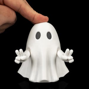 Peut inclure: Une figurine de fantôme blanche imprimée en 3D avec une tête arrondie lisse et deux yeux ovales noirs. Le fantôme possède deux bras articulés étendus vers l'extérieur, chaque main formant un signe de paix, avec des lignes de texture visibles.