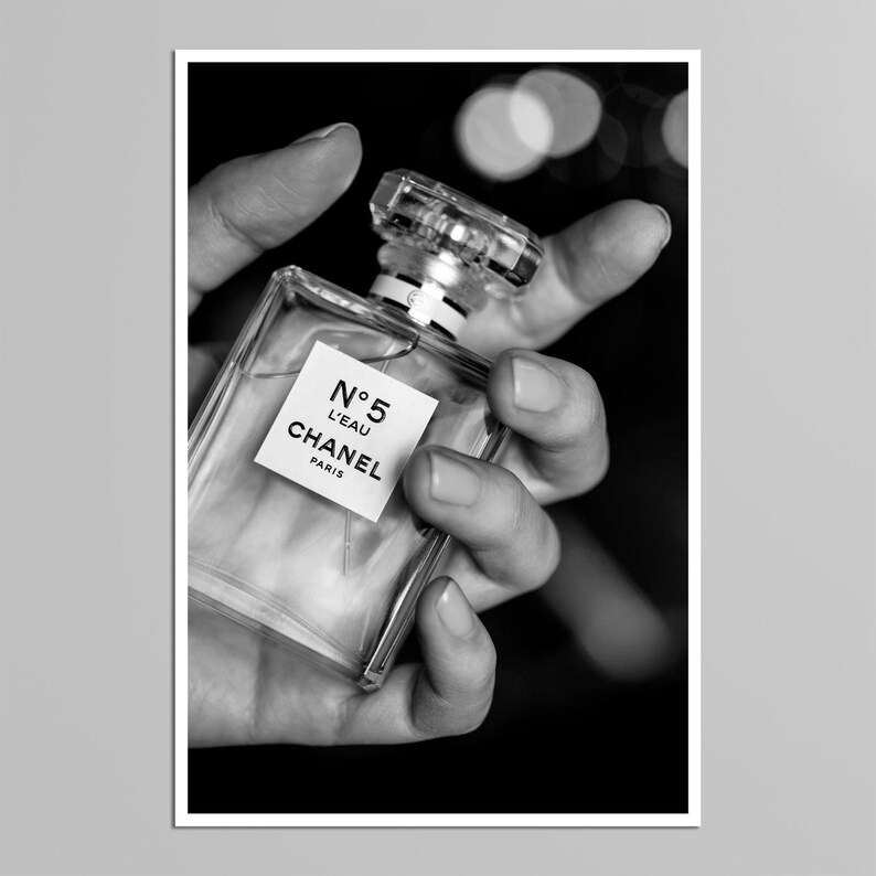 Puede incluir: Un frasco de perfume rectangular de vidrio transparente con un l&iacute;quido incoloro en su interior. El frasco presenta una etiqueta cuadrada blanca con el texto negro N&deg;5 L'EAU CHANEL PARIS. Tiene un tap&oacute;n de vidrio facetado y un cuello plateado.