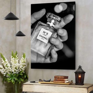 Puede incluir: Una impresi&oacute;n de arte mural en blanco y negro que muestra un frasco de perfume de vidrio transparente con una etiqueta cuadrada que dice N&deg;5 L'EAU CHANEL PARIS. El frasco se sostiene en una mano sobre un fondo oscuro con luces desenfocadas.