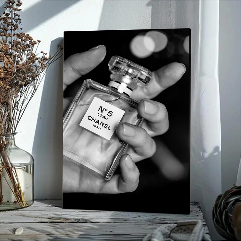 Puede incluir: Un cuadro rectangular en blanco y negro que muestra un frasco de perfume de cristal transparente con una etiqueta cuadrada que dice N&deg;5 L'EAU CHANEL PARIS. El frasco es sostenido por una mano sobre un fondo oscuro con suaves luces desenfocadas.