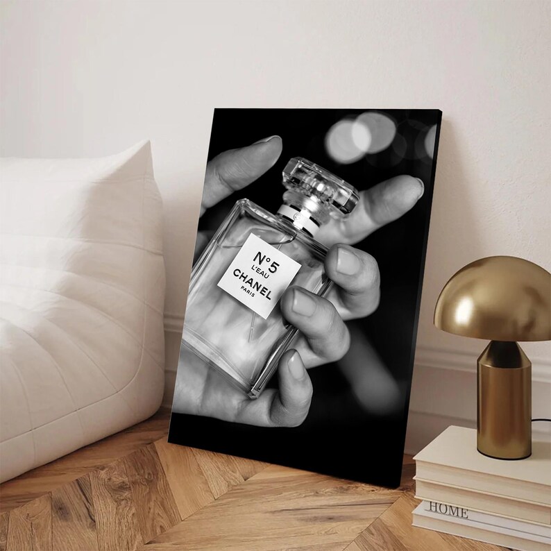 Puede incluir: Una impresi&oacute;n de arte mural rectangular en blanco y negro que muestra una mano sosteniendo un frasco de perfume de vidrio transparente. La etiqueta del frasco muestra claramente el texto N&deg;5 L'EAU CHANEL PARIS. El fondo es oscuro con luz bokeh.