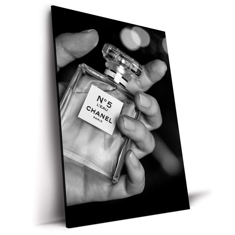 Puede incluir: Un frasco de perfume de cristal transparente con forma cuadrada y tap&oacute;n transl&uacute;cido. La etiqueta presenta el texto N&deg;5 L'EAU CHANEL PARIS en letras negras. El frasco aparece en una fotograf&iacute;a en blanco y negro de alto contraste.