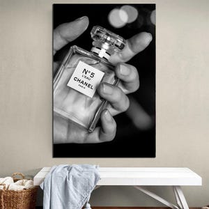 Puede incluir: Una fotograf&iacute;a en blanco y negro de un frasco de perfume de vidrio transparente con un tap&oacute;n cuadrado. La etiqueta en la parte frontal del frasco presenta el texto N&deg;5 L'EAU CHANEL PARIS en letras negras. El frasco se sostiene en una mano sobre un fondo oscuro.