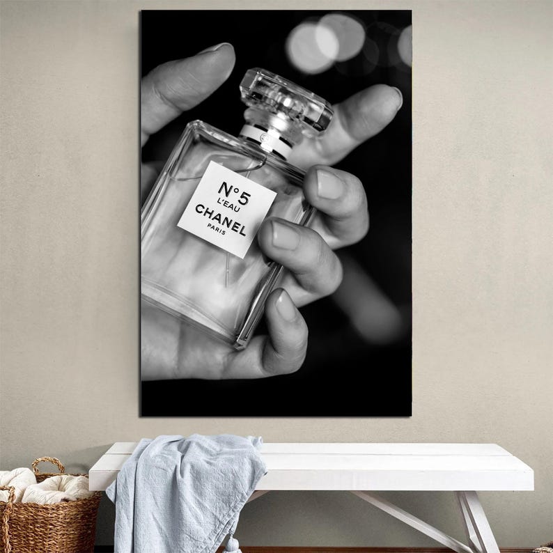 Puede incluir: Una fotograf&iacute;a en blanco y negro de un frasco de perfume de vidrio transparente con un tap&oacute;n cuadrado. La etiqueta en la parte frontal del frasco presenta el texto N&deg;5 L'EAU CHANEL PARIS en letras negras. El frasco se sostiene en una mano sobre un fondo oscuro.
