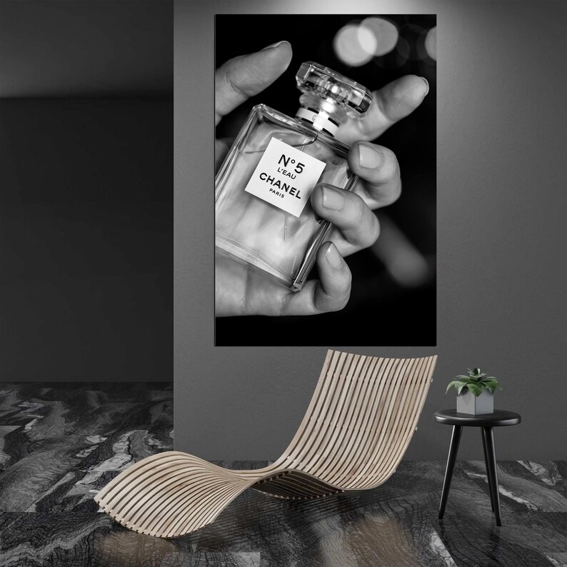 Puede incluir: Un cuadro rectangular de pared con una fotograf&iacute;a en blanco y negro de un frasco de perfume de cristal transparente con la etiqueta N&deg;5 L'EAU CHANEL PARIS, sostenido por una mano. La obra se exhibe sobre una tumbona de madera curvada.