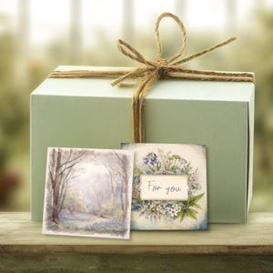 Puede incluir: Una caja de regalo verde salvia atada con cuerda, con dos tarjetas pequeñas. Una tarjeta muestra una escena de bosque, la otra tiene un diseño floral y las palabras "For you". La caja está sobre una superficie de madera.