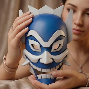 May include: A blue and white mask with a fierce expression, held by a person. The mask features a stylized design with sharp angles and a wide, toothy grin. The mask's color scheme is primarily blue and white.
