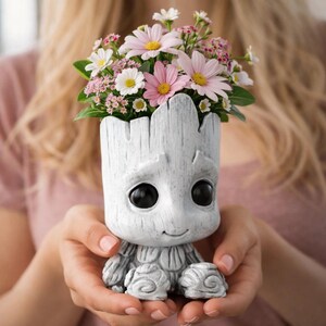 May include: A gray, Groot-shaped planter holding a bouquet of pink, white, and yellow flowers. The planter has large black eyes and a smiling expression. The flowers are a mix of daisies and other small blooms.