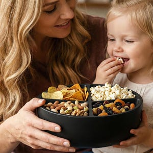 Puede incluir: Un cuenco negro dividido en secciones, lleno de aperitivos, incluyendo patatas fritas, palomitas de maíz, frutos secos y fruta deshidratada. Una mujer y un niño pequeño sostienen el cuenco. El niño está comiendo palomitas.
