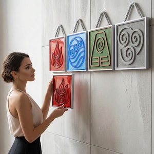 Puede incluir: Una mujer sostiene una obra de arte mural enmarcada en rojo con un diseño de llama. Otras cuatro obras de arte murales enmarcadas cuelgan de la pared, cada una con un símbolo elemental diferente: fuego, agua, tierra y aire. Los marcos son plateados con colgadores de cuerda.