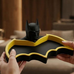 May include: A black and yellow Batman-themed tray. The tray is shaped like the Batman logo, with a black interior and a yellow border. A black Batman bust is centered on the tray.