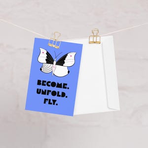 Könnte beinhalten: Eine blaue Grußkarte mit einer schwarz-weißen Schmetterlingsillustration mit rosa Akzenten und dem Text BECOME. UNFOLD. FLY. in fetten schwarzen Buchstaben. Die Karte ist mit einem goldenen Metallclip an einer Schnur neben einem weißen Umschlag befestigt.
