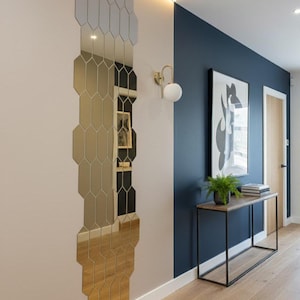 Op de afbeelding: Een moderne hal met een geometrische spiegel, een console en een ingelijst kunstwerk. De muren zijn geschilderd in contrasterende kleuren: lichtbeige en marineblauw. Een gouden wandlamp met een witte bol zorgt voor verlichting. Een kleine plant en boeken sieren de tafel.