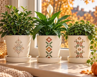 May include: Three off-white ceramic plant pots with matching saucers, each featuring a unique cross-stitch style geometric pattern in muted autumn colors. The pots contain green foliage, including a peace lily with white spathe flowers.