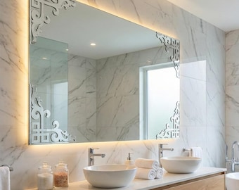 May include: A large rectangular bathroom mirror with decorative corner accents and integrated lighting. The mirror is mounted above a wooden vanity with two white vessel sinks. The background features marble-patterned walls and a window.
