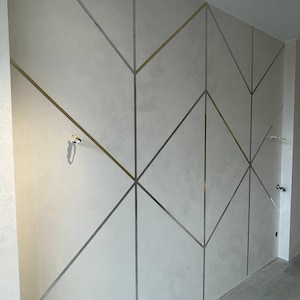 May include: A wall featuring a light gray textured finish divided by a geometric pattern of thin, metallic gold and silver strips. The design consists of intersecting lines forming various diamond and triangular shapes across the flat surface.