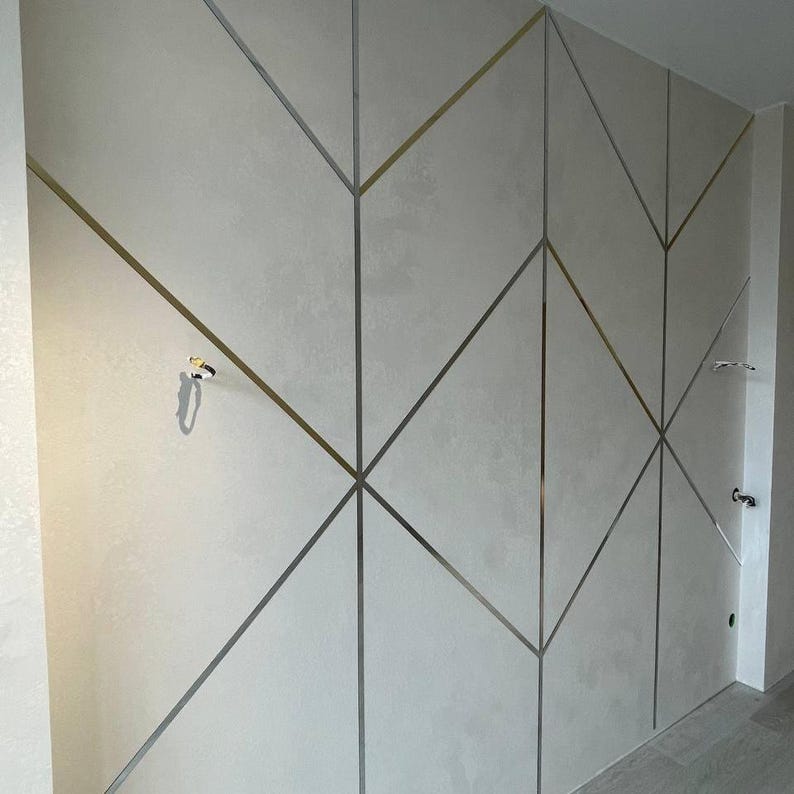 May include: A wall featuring a light gray textured finish divided by a geometric pattern of thin, metallic gold and silver strips. The design consists of intersecting lines forming various diamond and triangular shapes across the flat surface.