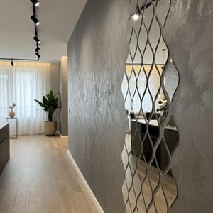 Puede incluir: Un pasillo interior moderno con un espejo de pared decorativo. El espejo está compuesto por múltiples piezas onduladas interconectadas. Las paredes son de un gris texturizado y el suelo es de madera clara. También se ve una planta en maceta e iluminación de riel.