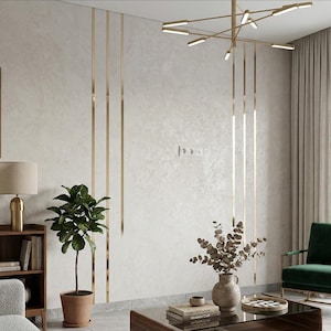 May include: A wall featuring light gray textured plaster with vertical gold metal inlay strips, a modern gold chandelier, a potted green plant, a wooden bookshelf with books, a vase with dried branches, and a dark green armchair.