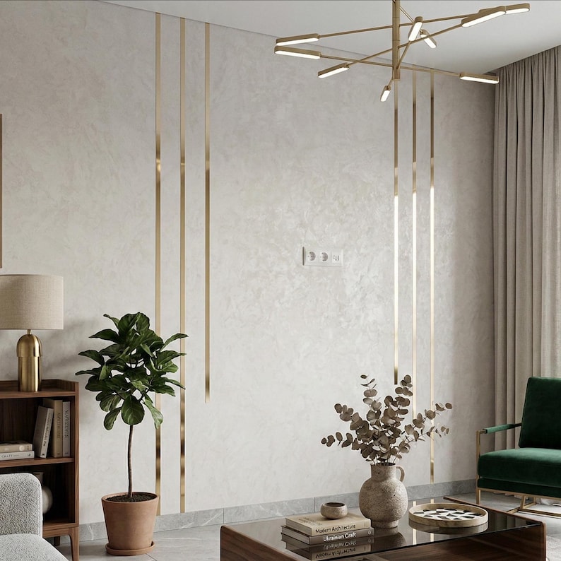 May include: A wall featuring light gray textured plaster with vertical gold metal inlay strips, a modern gold chandelier, a potted green plant, a wooden bookshelf with books, a vase with dried branches, and a dark green armchair.