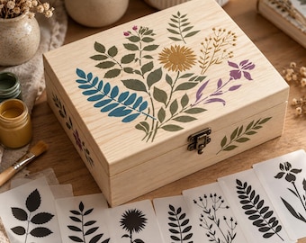 May include: A rectangular light-colored wooden box with a metal latch, featuring a painted botanical design of a sunflower, leaves, and sprigs in blue, green, gold, and purple. Several black and white floral stencils are arranged in front.