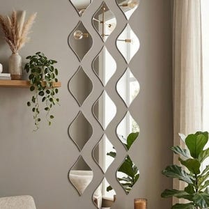 Könnte beinhalten: Eine Anordnung dekorativer Wandspiegel in einem vertikalen, wellenförmigen Muster. Jeder Spiegel hat eine einzigartige, längliche Form mit einer reflektierenden Oberfläche. Die Spiegel sind an einer neutralfarbenen Wand angebracht und reflektieren den Raum.