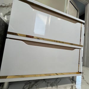 May include: A two-drawer vanity cabinet featuring a glossy white finish on the drawer fronts, light wood grain accents, and horizontal gold-toned metal trim. The cabinet is supported by cylindrical silver-toned legs and has a rectangular shape.