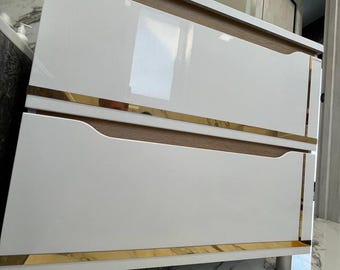 May include: A two-drawer vanity cabinet featuring a glossy white finish on the drawer fronts, light wood grain accents, and horizontal gold-toned metal trim. The cabinet is supported by cylindrical silver-toned legs and has a rectangular shape.