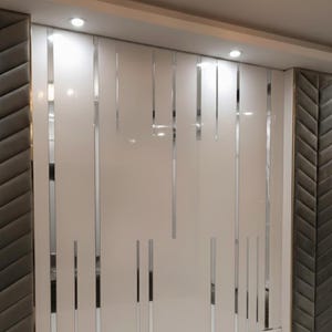 May include: A white wall panel with vertical silver strips and recessed lighting. The panel is flanked by sections of gray, textured material with a chevron pattern. The design is modern and minimalist.