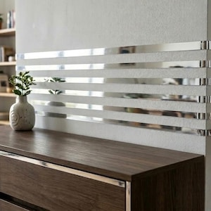 May include: A series of horizontal, reflective silver mirror strips applied to a textured white wall. The strips create a linear pattern that reflects the surrounding room. A small, light-colored ceramic vase with a plant sits nearby.