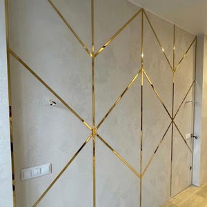 May include: A wall paneling system featuring light gray, textured surfaces divided by thin, reflective gold-colored metal strips arranged in a geometric pattern of intersecting lines and diamonds, including a white light switch plate.