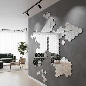 Puede incluir: Un espejo de pared compuesto por numerosas baldosas hexagonales, dispuestas en un diseño similar a un mapa. El espejo refleja el interior de la habitación, incluyendo un sofá verde, una silla de madera y una planta en maceta, contra una pared gris.