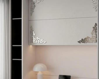 May include: A set of four silver-toned decorative corner appliqués with intricate scrollwork patterns, mounted on the corners of a white cabinet door. The metallic accents feature symmetrical, ornate designs that contrast against the flat surface.