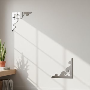 Könnte beinhalten: Zwei dekorative silberfarbene Metall-Winkelhalterungen mit einem stilisierten, geschwungenen Design sind an einer weißen Wand montiert. Die Halterungen sind in gegenüberliegenden Ecken positioniert und zeigen ihre symmetrische Form und Eignung für Regale.